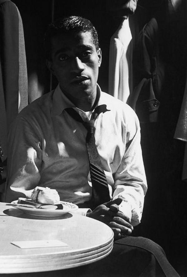 A black-and-white photo shows a man sitting at a table with a dessert plate in front of him. He wears a white shirt, a loosened tie, and an expression of contemplation. The background is dimly lit with clothing items hanging.