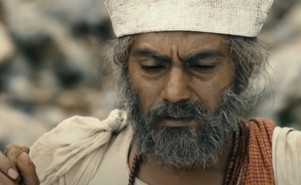 A man with a gray beard and mustache is wearing a white cloth headpiece and traditional attire, including a beaded necklace and a shoulder drape. He appears deep in thought, with a serious expression, and is holding a small object in his right hand.