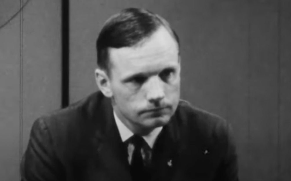 Black-and-white photo of a man with short hair wearing a suit and tie, looking forward with a serious expression. The background is plain and appears to be indoors.