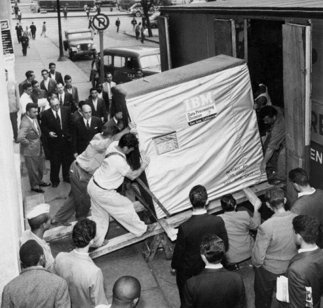 Workers unload a large, boxed IBM data processing system from a truck while bystanders watch, in a bustling, mid-20th-century urban street scene.