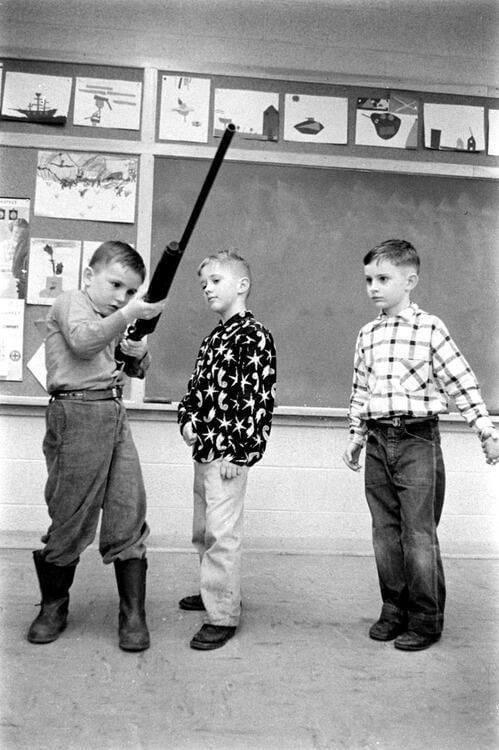 Black-and-white photo of three young boys in a classroom; the boy on the left is holding and looking down the barrel of a long rifle, the middle boy watches attentively, and the boy on the right observes with a casual stance. Classroom posters are visible in the background.