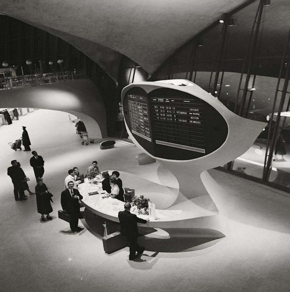 A black and white image of a mid-20th-century airport terminal with a large information display overhead. Several people are gathered around a circular information desk. The terminal features futuristic, curved architectural elements and large windows.