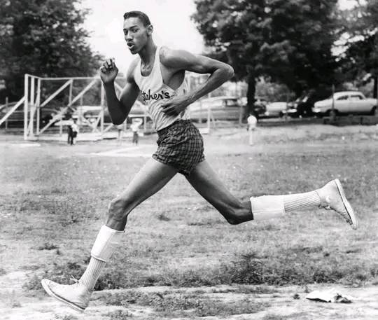 Black-and-white photo of a tall athlete running outdoors. He wears a tank top with text, shorts, and knee-high socks. Trees, parked cars, and a few people are visible in the background. He appears focused and determined as he strides forward.