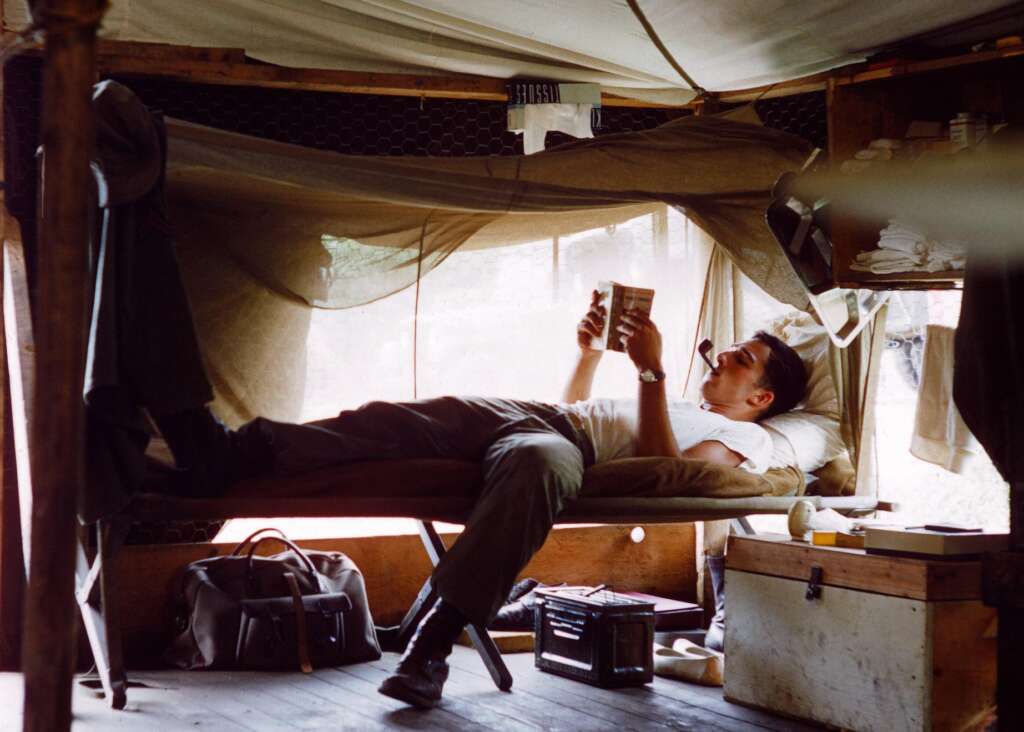 A man in casual attire lies on a makeshift bed inside a tent, reading a book. The tent is furnished modestly with a suitcase, crate, and hanging fabrics. Sunlight filters softly through the tent's window, casting a warm, relaxed ambiance.