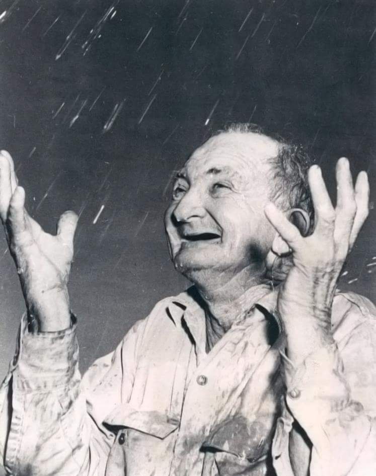 An elderly man looks up joyfully with his arms raised as rain falls. He wears a wet shirt, and the background is dark, highlighting the rain. His expression is one of happiness and relief, capturing a moment of pure enjoyment in the rain.