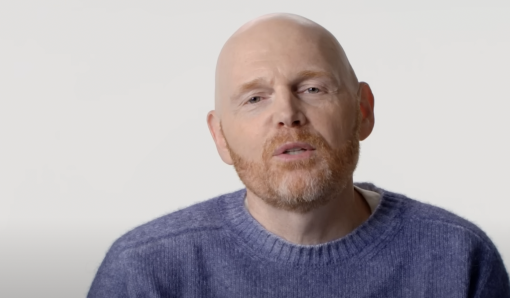 A bald man with a reddish beard is wearing a blue sweater and is speaking against a plain white background.