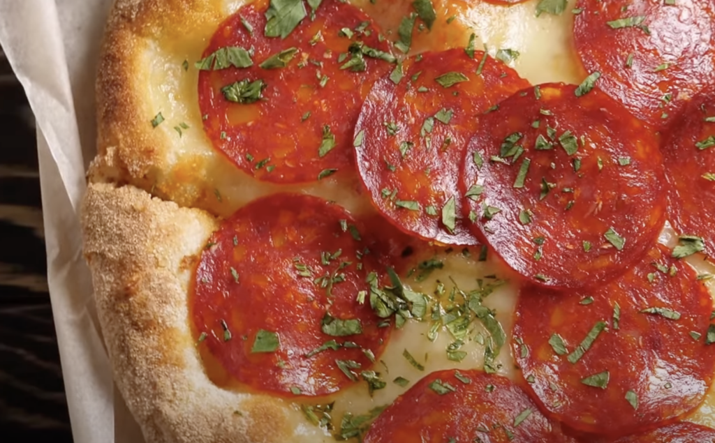 A close-up view of a pepperoni pizza. The pizza has a golden-brown crust, topped with melted cheese, pepperoni slices, and a sprinkling of fresh parsley. The pizza is presented on a piece of parchment paper.