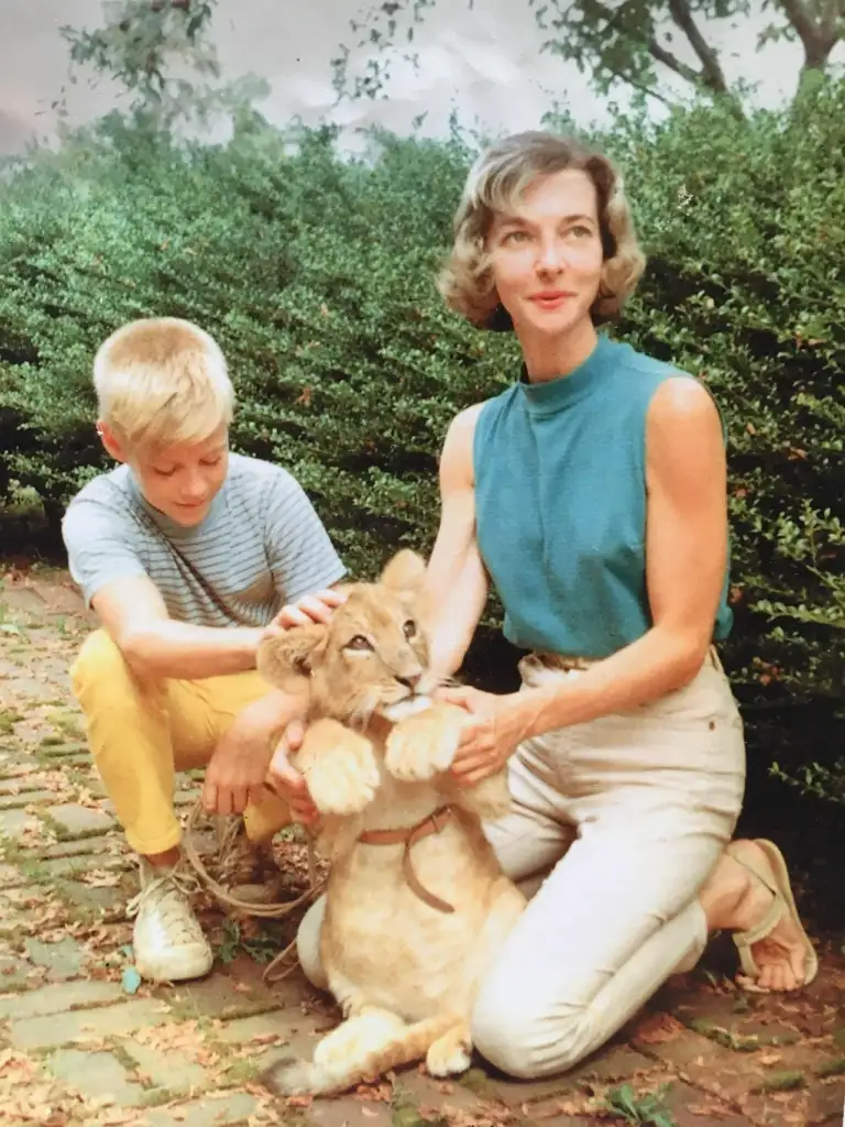 A woman kneels on the ground next to a lion cub, holding its paws, while a young boy wearing yellow pants and a striped shirt kneels beside them, petting the cub. They are outdoors, surrounded by greenery.