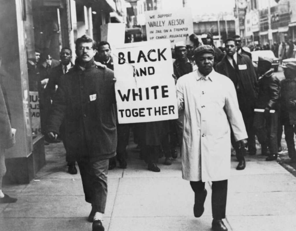 A black-and-white photo depicting a civil rights march with a diverse group of demonstrators. Two men in front hold signs reading "BLACK AND WHITE TOGETHER". They walk along a city street, with more demonstrators and buildings visible in the background.