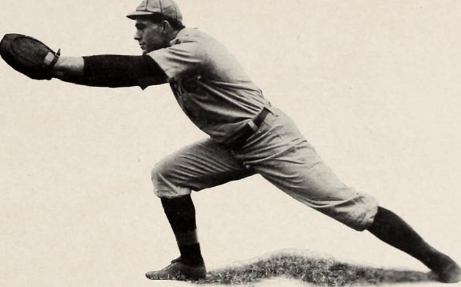 A vintage photograph shows a baseball player dressed in an old-fashioned uniform. He is leaning forward with one knee bent, stretching out his gloved hand to catch a ball. The image has a nostalgic feel with its black-and-white tones.