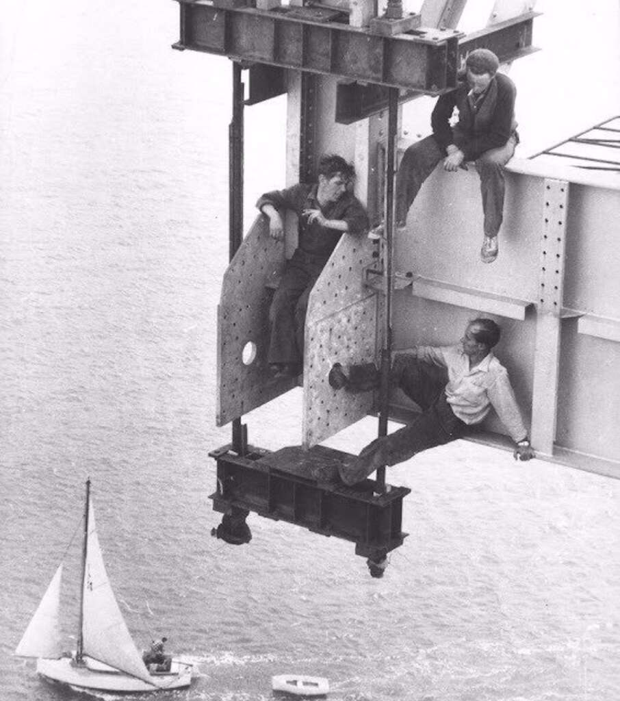 Three construction workers sitting casually on a steel beam high above water, with one worker dangling his legs and another leaning back. Below them, a small sailboat navigates the water. The scene depicts a high-risk environment and vertigo-inducing heights.