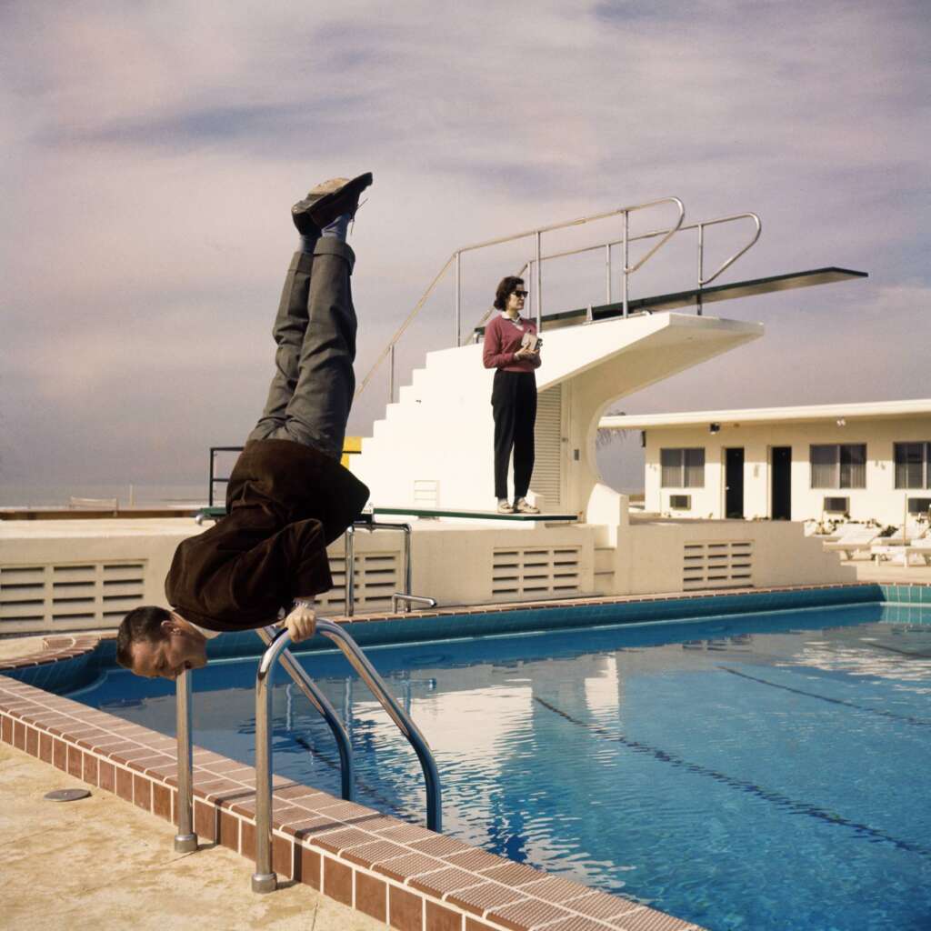 A person in a brown jacket performs a handstand at the edge of an outdoor swimming pool while gripping the metal ladder. In the background, another person stands on a diving board holding a magazine. The sky is partly cloudy with a soft, ambient light.