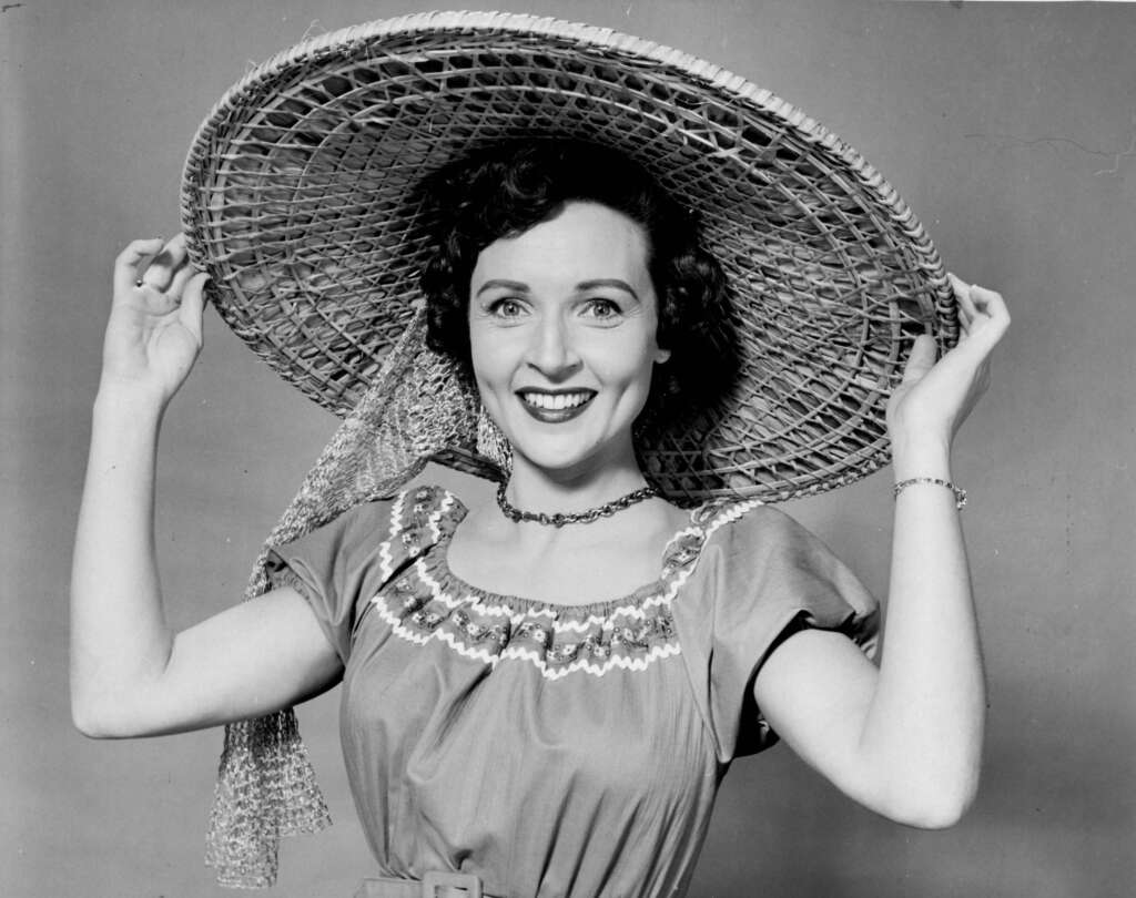 A woman wearing a large, wide-brimmed hat smiles brightly at the camera. She has curly hair and is dressed in a belted dress with ruffled, embroidered detailing at the neckline. She holds the brim of her hat with both hands. The background is plain.