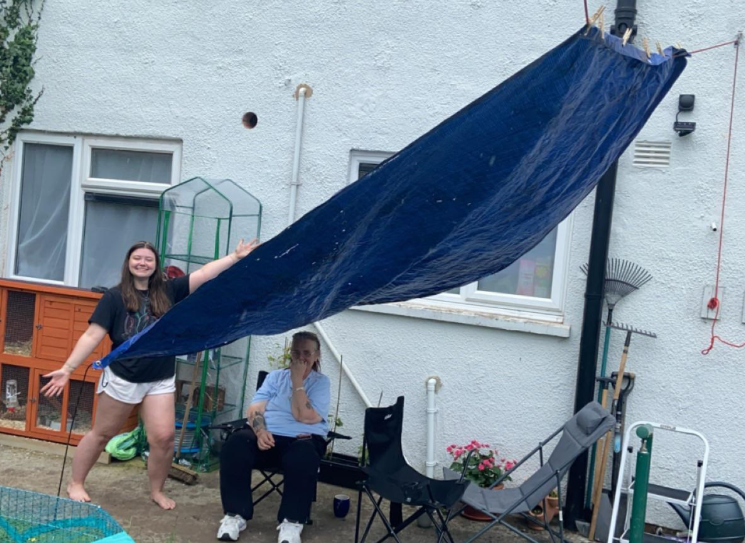 Two individuals are in an outdoor yard. One person stands with arms outstretched next to a makeshift blue tarp canopy, while the other sits on a chair underneath it, appearing thoughtful. Surrounding them are various garden tools and a plant.