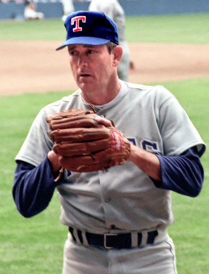 A baseball player in a grey uniform with "RANGERS" written on the front, wearing a blue cap with a red and white "T", holds a glove in both hands while on a baseball field. The background shows the grass outfield and part of the infield dirt.