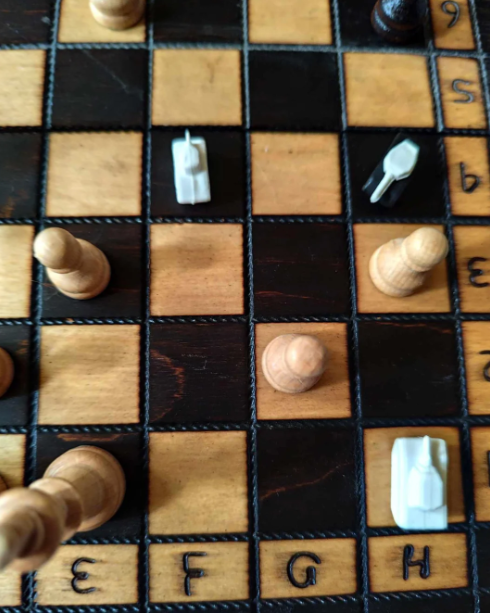 An overhead view of a chessboard with wooden and plastic pieces. The board is not set up for a standard game, featuring unconventional pieces that resemble vehicles or buildings, positioned on various squares. The board is labeled with coordinates along the edges.