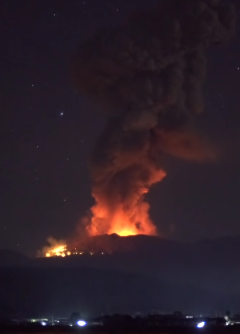 Fiery eruption of a volcano at night, with bright orange lava and smoke ascending into the dark sky, illuminated by the burning intensity of the eruption. Distant lights from a settlement are visible at the bottom of the image.