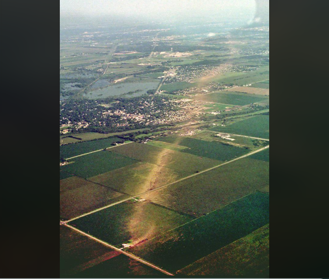 Aerial view of a landscape showing a tornado's path through a rural area. The tornado's trail is visible as a strip of damage cutting through green fields and a small town, with clear skies above.