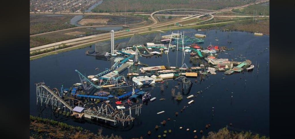 Aerial view of an abandoned, flood-damaged amusement park with submerged rides, buildings, and pathways. Surrounding areas also appear waterlogged, with a highway and sparse vegetation visible in the background. The scene appears desolate and eerie.