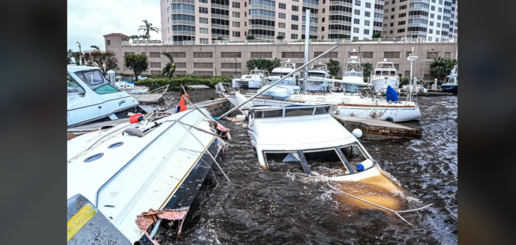 Boats are damaged and partially submerged in rough waters at a marina, with some tipped over and others heavily leaning to one side. High-rise buildings stand in the background, and broken debris is scattered throughout the scene.