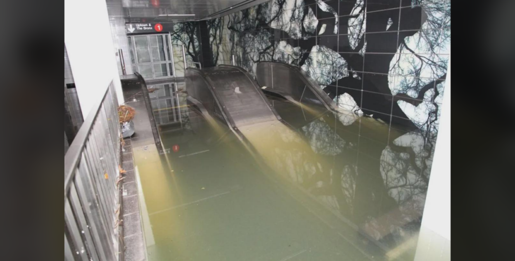 A flooded subway station with murky water submerging the escalators. The walls feature a dark tile mural with tree branch designs. A "No Smoking" sign is visible above the waterlogged entrance.