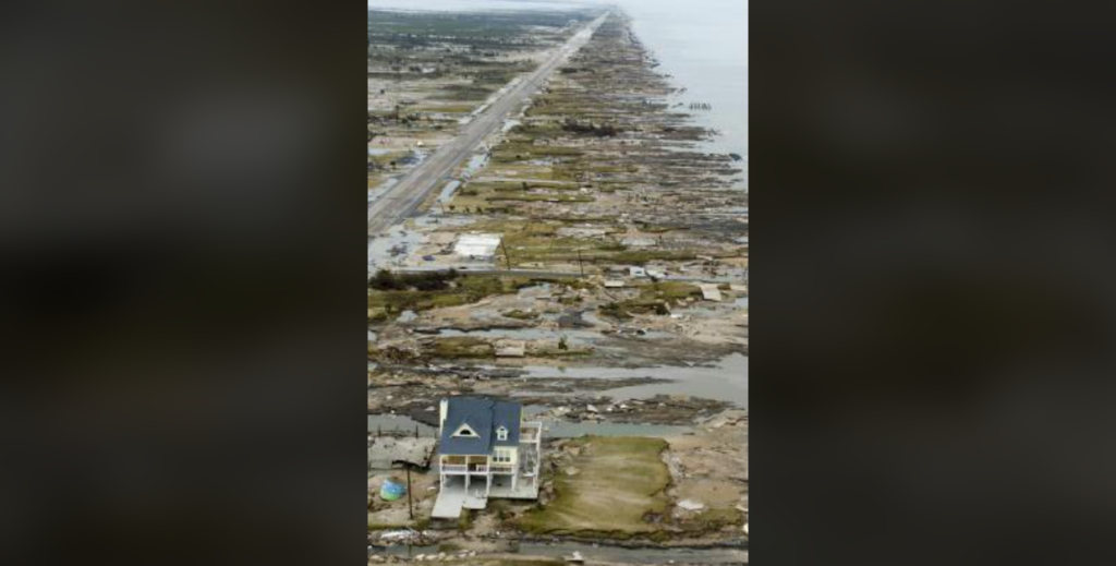 Aerial view of a coastal area with severe storm damage. A single house remains standing near the foreground, surrounded by debris-strewn land. The road runs parallel to the shoreline, with additional wreckage extending into the distance.