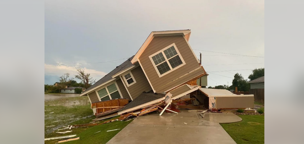 A two-story house has completely collapsed, with the upper floor leaning sharply to the side. Debris from the house is scattered around, indicating severe structural damage. The surrounding area is wet, suggesting recent heavy rainfall or flooding.