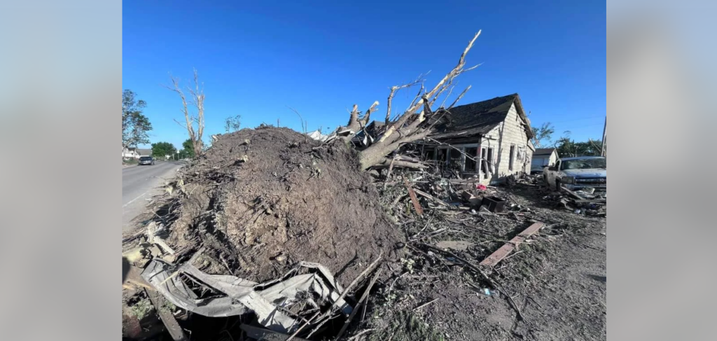 A large tree has fallen on a house, causing significant damage. Debris and broken branches are scattered around, and the house is partially collapsed. The sky is clear and blue, indicating a likely recent storm. A vehicle is visible in the background.