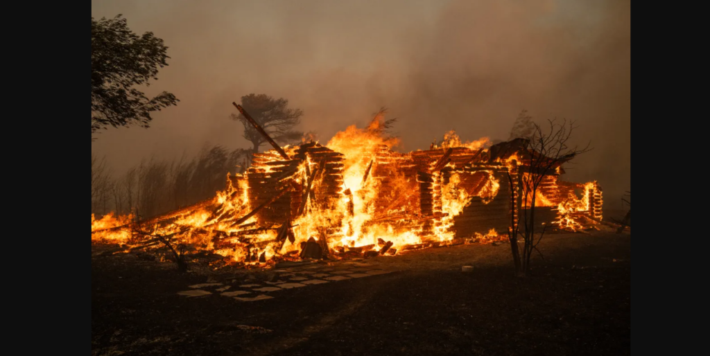 A large fire consumes a wooden structure, with flames engulfing the entire building. The surrounding area is dark and smoky, with charred trees and debris visible. The intense blaze emits a bright orange glow against the dark, smoky background.