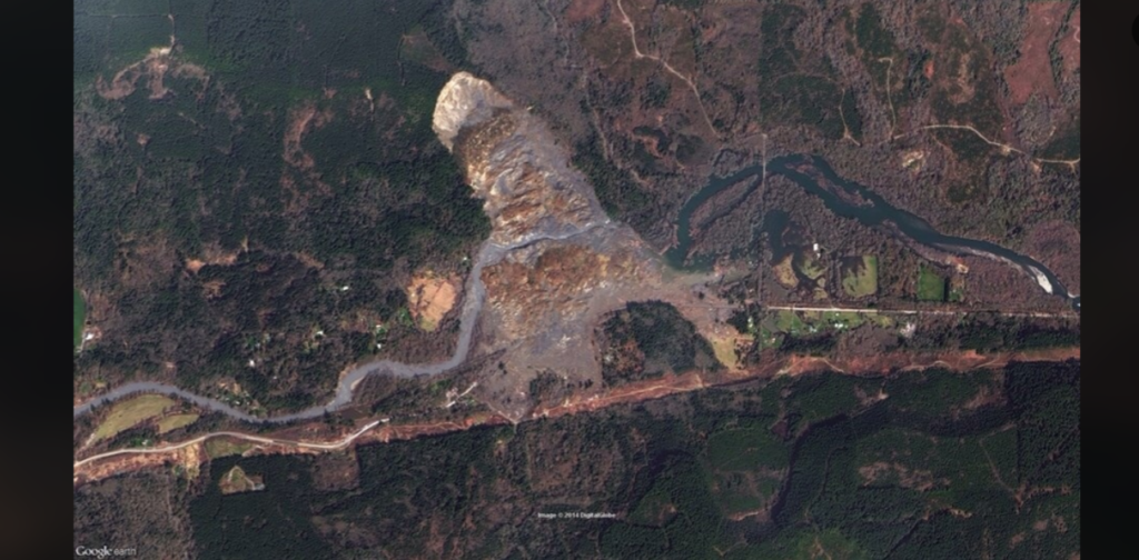 A satellite image shows a large landslide area with displaced earth laid out in a fan shape. The affected expanse includes a river diverting around the debris, nearby residences, and roads. Surrounding areas are covered in dense green vegetation.