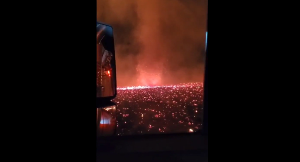 View from a vehicle showing the aftermath of a large fire at night. The landscape is covered in smoldering embers and glowing flames under a dense, smoky sky, creating an eerie and apocalyptic scene. Vehicle mirrors are visible on the left side.