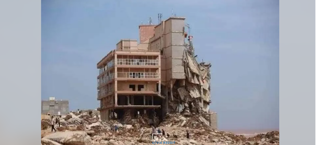 A partially collapsed multi-story building stands amidst rubble and debris against a clear blue sky. The right side of the structure is severely damaged, exposing the interior and appearing unstable, contrasting with a relatively intact left side. Nearby, smaller structures are also affected by the destruction.