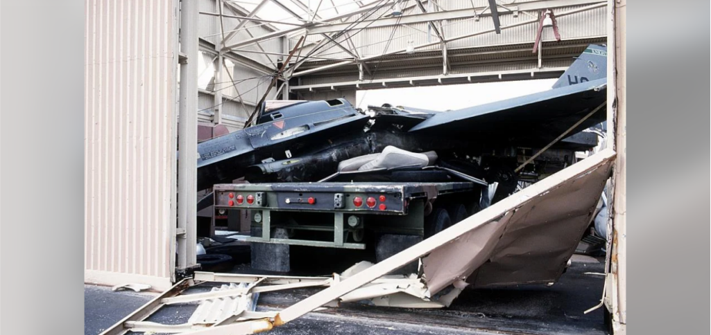 An aircraft with visible damage appears to have crashed into a building, causing significant structural damage to the corrugated metal and frame. The tail section is raised, indicating the impact. The surrounding area is strewn with debris.