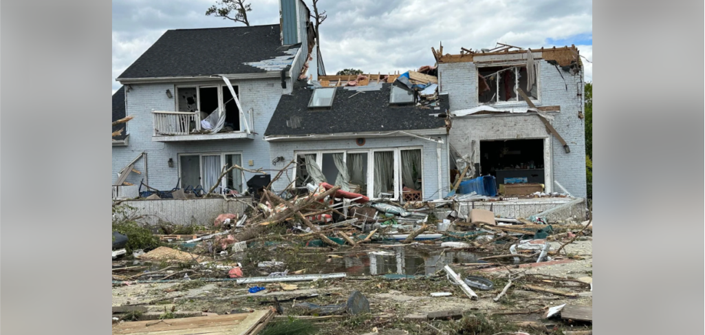 A two-story house heavily damaged by a severe storm or tornado. The roof is partially torn off, windows are shattered, and debris is scattered across the yard, including tree branches and household items. The sky is cloudy, suggesting a recently passed storm.