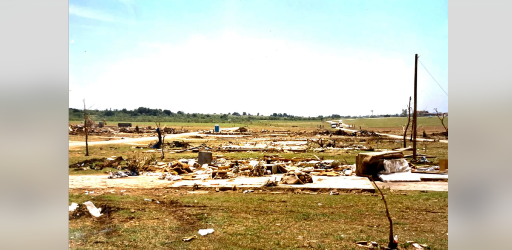 A field strewn with debris and remnants of structures following a natural disaster, possibly a tornado. Scattered trees and utility poles remain standing amidst the devastation, while the horizon shows a sparse, flat landscape under a clear sky.