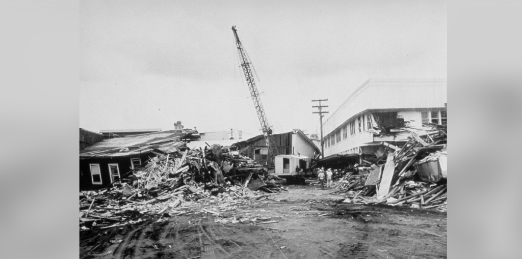 Black and white photo of a street filled with debris and destroyed buildings. A crane is visible in the background amidst the ruins. The scene suggests extensive damage, possibly from a natural disaster. Buildings on both sides appear heavily damaged and collapsed.