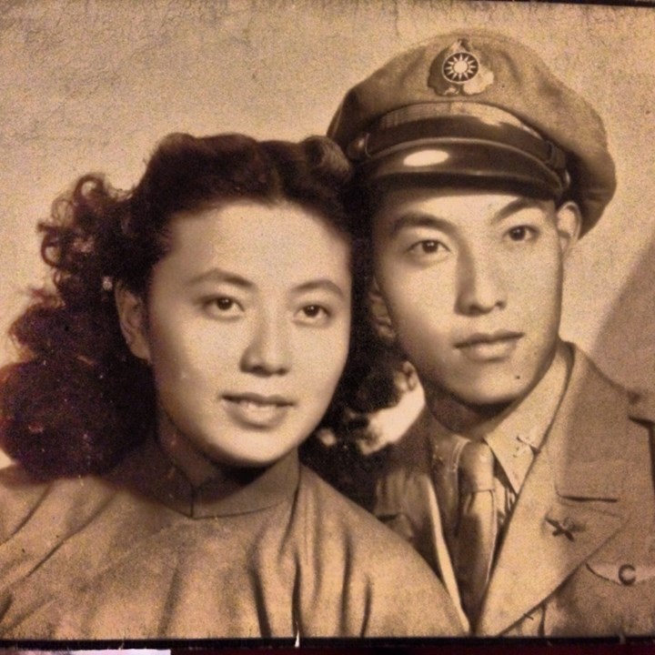 A vintage black-and-white photo of a couple posing together. The woman has curly hair and wears a traditional outfit, while the man, wearing a military uniform with a peaked cap adorned with insignia, looks straight ahead. The image has a worn, nostalgic feel.