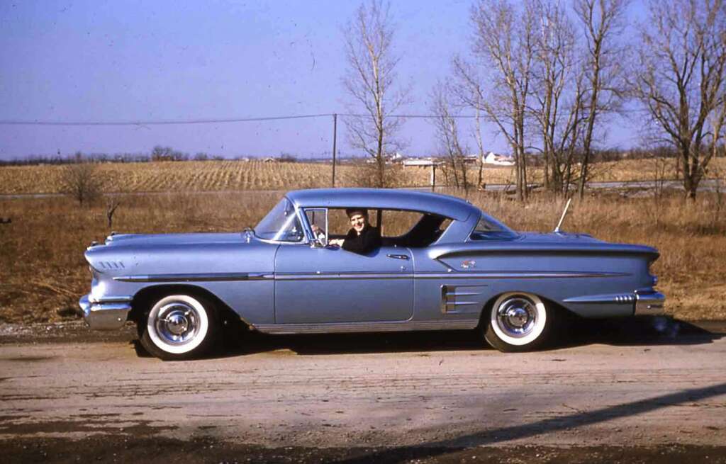 A man sits in the driver's seat of a vintage blue 1958 Chevrolet Impala parked on a rural road. The scenery features a field with bare trees under a clear sky, capturing an autumn or early winter ambiance.