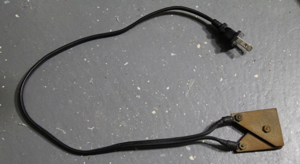 A black cable with a two-prong plug on one end and a rusty, triangular metal piece with two wires attached on the other end lies on a gray speckled surface.