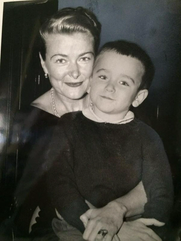 A black-and-white photo of a smiling woman with 1940s hairstyle and pearl necklace, holding a young boy on her lap. The boy has short dark hair and is wearing a sweater. The woman looks content and the boy looks calmly at the camera.