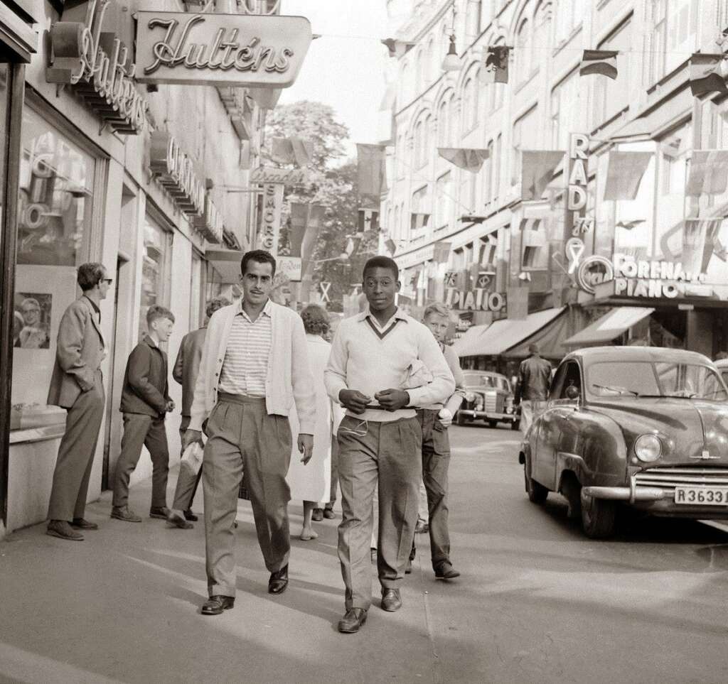 Two men walk down a bustling city street lined with various shops, including one with a sign that reads "Julien's." Pedestrians are visible on the sidewalk, and a vintage car is parked on the road. The scene appears to be from a past era, possibly the mid-20th century.