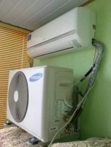 An indoor air conditioning unit installed on a wall with its corresponding outdoor compressor unit positioned on a shelf directly below it. Both units are connected by insulated refrigerant pipes.