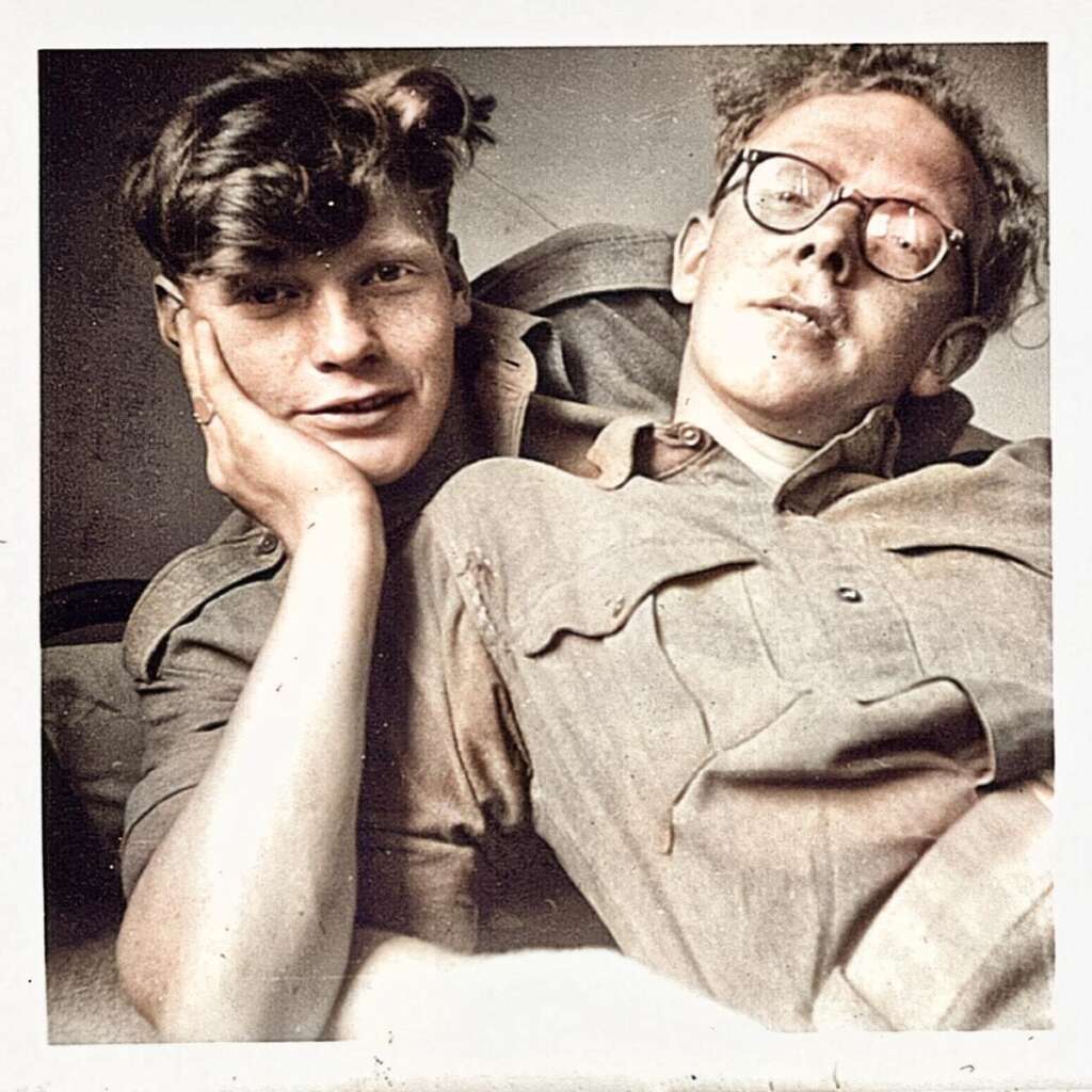 Two individuals in military uniforms relaxing together. The person on the left has tousled hair, resting a cheek on their hand, while the person on the right, wearing glasses, reclines comfortably. The image is vintage with a sepia tone effect.