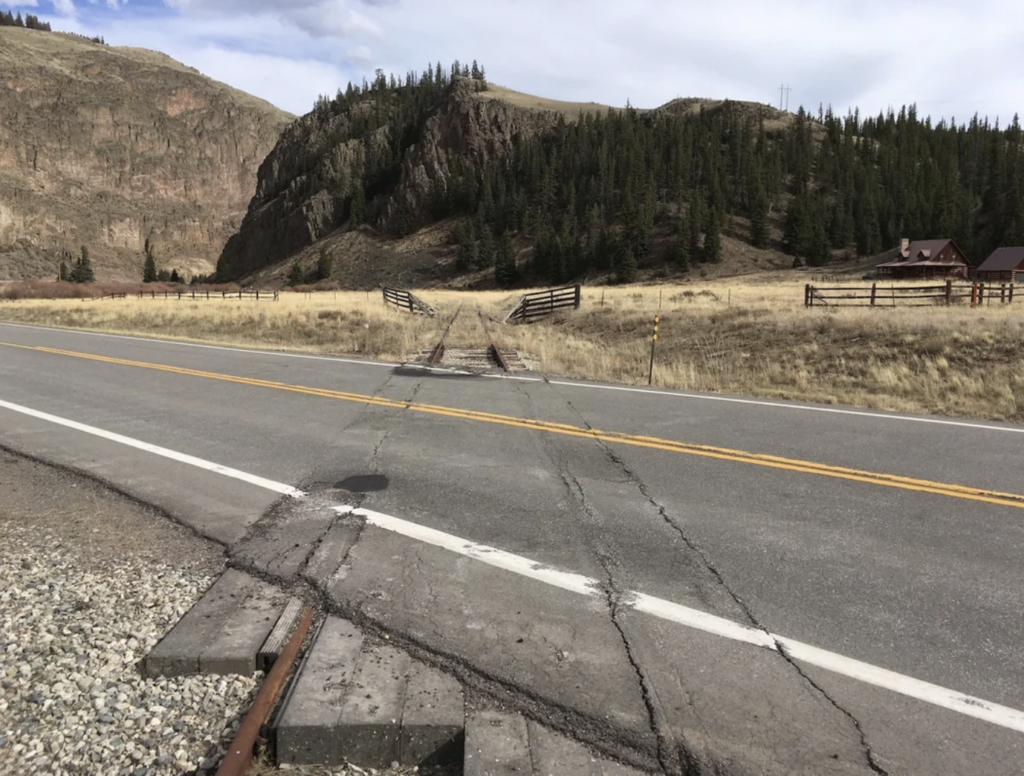 An empty two-lane road meets a disused railroad track in a rural area. Distant hills covered in evergreen trees and a clear sky serve as the backdrop. Fencing lines the road and a house is visible on the right side.