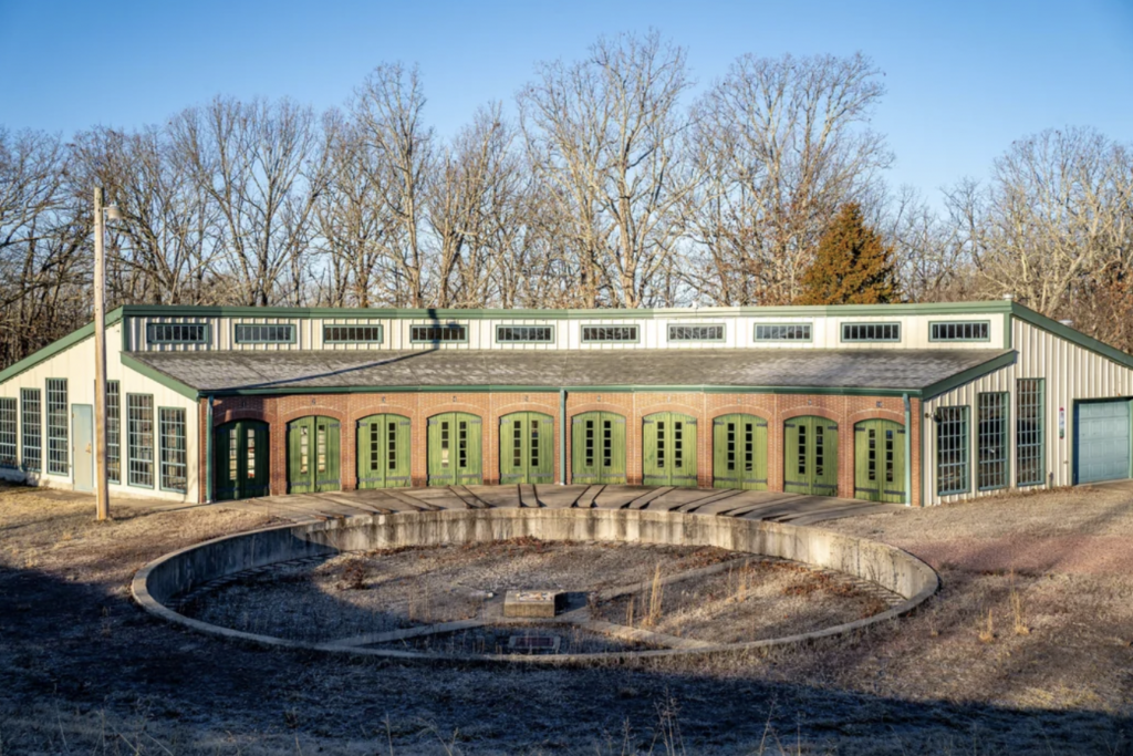 A roundhouse with multiple green doors stands in a clearing, encircled by tall, leafless trees under a clear blue sky. The structure shows a semi-circular track in the foreground and an open area in front, with weathered ground and sparse vegetation.