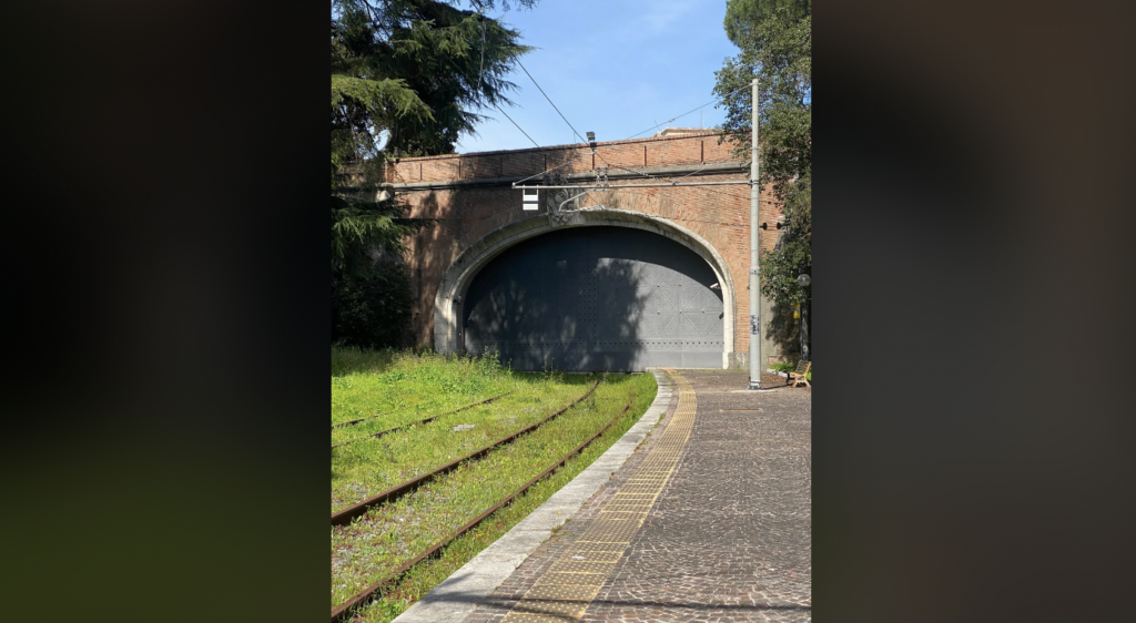 A tunnel entrance with a large arched opening made of red bricks. The tunnel is closed with a large metal door. Tram tracks lead into the tunnel, and there is greenery on one side and a cobblestone path on the other. Trees and blue sky are visible above.