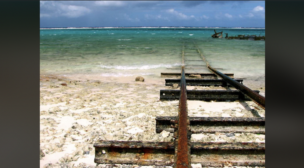 Old, rusty railway tracks lead directly into calm, turquoise ocean waters from a white, rocky shore. The tracks are partially submerged, disappearing into the sea. The sky is overcast, adding a grayish hue to the overall scene.