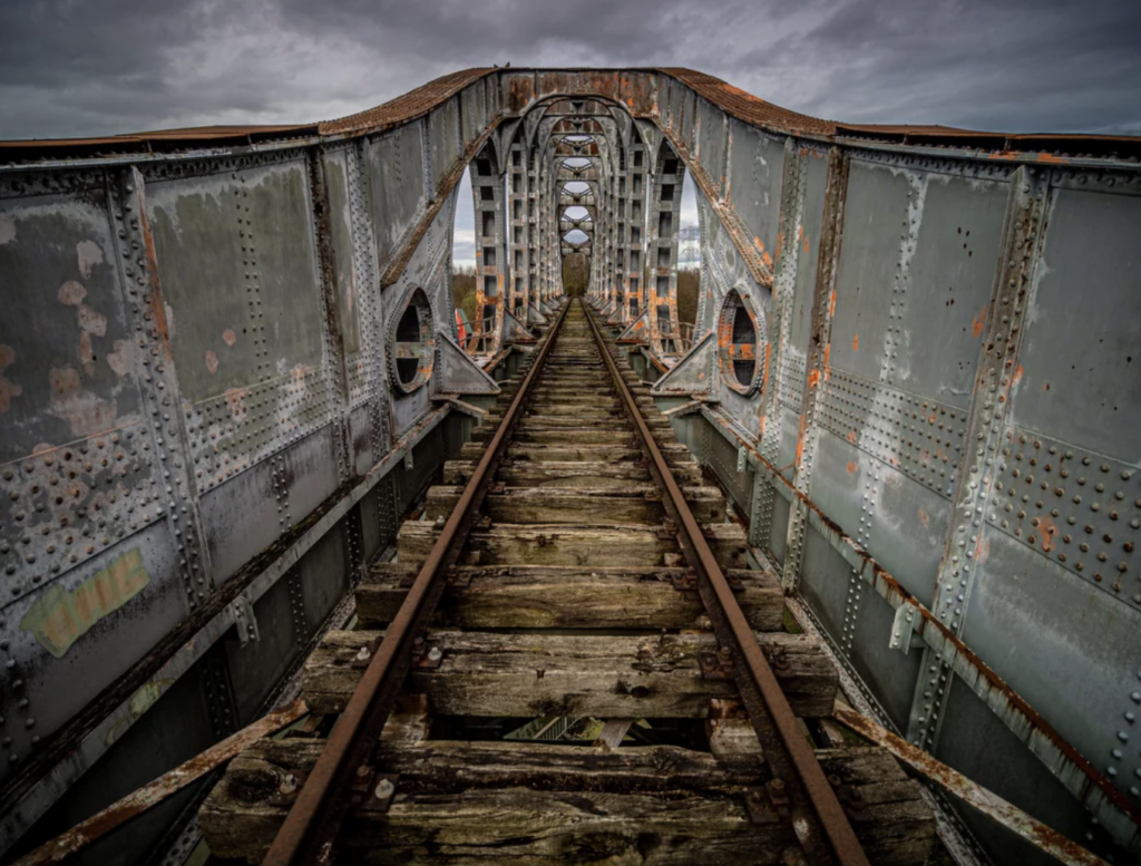 An old, rusty metal bridge with wooden railway tracks stretches into the distance under an overcast sky. The bridge features large, rectangular support beams and riveted panels, showing signs of decay with peeling paint and patches of corrosion.