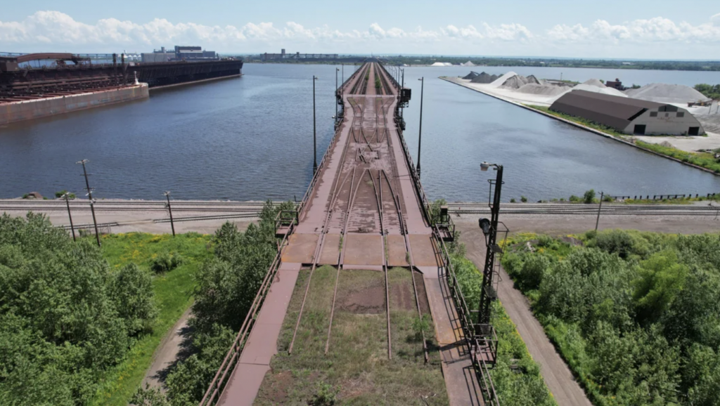 A long, rusty bridge extends over a body of water toward a distant industrial area. The bridge has multiple train tracks and overgrown vegetation in the center. On the right, there are piles of industrial materials, and on the left, large industrial buildings.