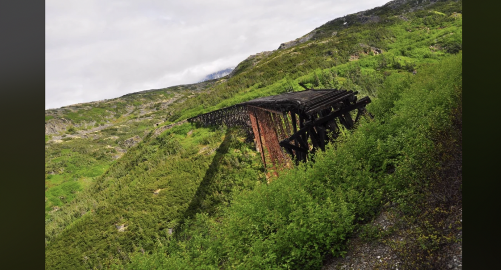 An old, abandoned wooden bridge stands in a lush, green mountainous landscape. The bridge is weathered, with some parts missing, and casts a long shadow on the dense foliage below. The sky is overcast, adding a sense of melancholy to the scene.